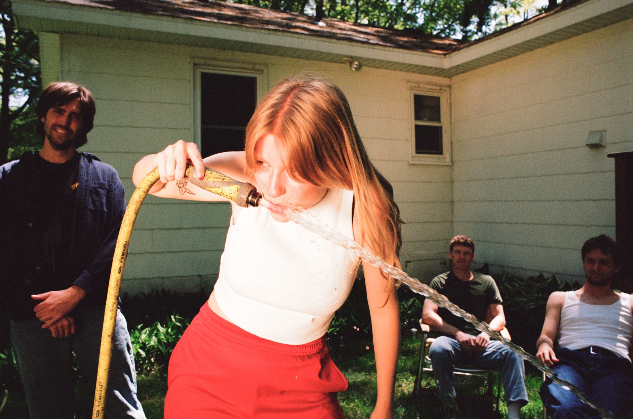 Slow Pulp band members gather around the lead singer as she drinks from a garden hose in the yard behind a house